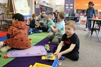 Students in TK engage in purposeful play in foreground while students listen to a book being read aloud in the background.