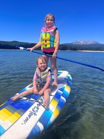 Two kids wearing life jackets and learning how to stand up paddleboard with their parents supervising nearby.